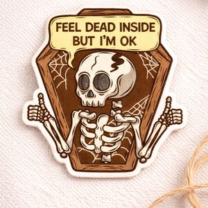May include: A sticker featuring a cartoon skeleton inside a coffin, giving a thumbs-up with both hands. The skeleton has a speech bubble that says "FEEL DEAD INSIDE BUT I'M OK". The sticker is brown, beige, and white.