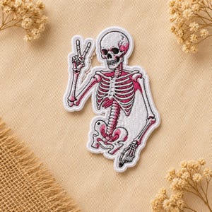 May include: Embroidered patch featuring a white skeleton with pink accents, giving a peace sign. The patch has a detailed design of the skeletal structure, including the skull, rib cage, and limbs. The patch is outlined in black.