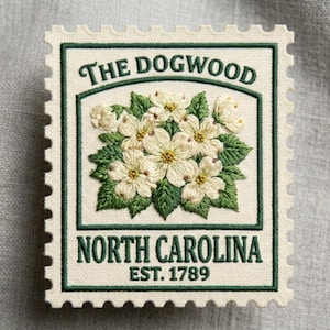 May include: Embroidered stamp featuring a dogwood flower design. The stamp has a green border and text that reads "THE DOGWOOD", "NORTH CAROLINA", and "EST. 1789". The flowers are white with yellow centers and green leaves.