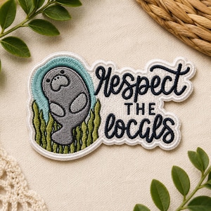 May include: Embroidered patch with a grey manatee in a teal and green aquatic scene. The patch reads "Respect the Locals" in a cursive font. The patch is bordered with white stitching.