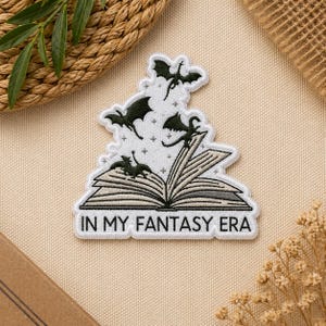 May include: Embroidered patch featuring an open book with dragons flying out. The patch is white with black and gray details, and the text "IN MY FANTASY ERA" is at the bottom. The patch is on a beige background.