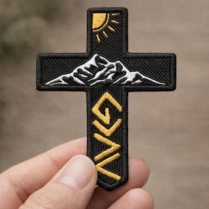 May include: Black embroidered cross patch featuring a mountain range, sun, and the letters "GNG" in gold. The patch is held against a blurred background, showcasing the detailed stitching and design.