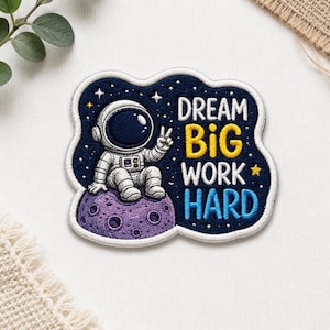 May include: Embroidered patch featuring an astronaut seated on a purple planet, against a dark blue starry background. The patch reads "DREAM BIG WORK HARD" in white, yellow, and blue lettering. The astronaut is wearing a white spacesuit and giving a peace sign.
