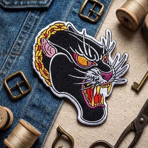 May include: Black panther embroidered patch with yellow, red, and white accents. The patch is on a denim jacket, surrounded by sewing supplies and metal buckles. The panther has an open mouth with visible teeth.