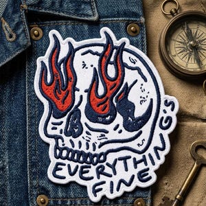 May include: Embroidered patch featuring a white skull with red flames for eyes and navy blue accents. The text "EVERYTHING FINE" is below the skull. The patch is on a denim jacket.