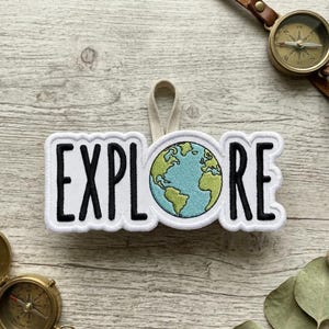 May include: A white embroidered patch with the word "EXPLORE" in black letters, featuring a globe in the center. The patch has a cream-colored loop for hanging. A compass and other travel-themed items are in the background.