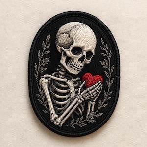 May include: Embroidered patch featuring a detailed skeleton holding a red heart. The skeleton is rendered in white thread against a black oval background, framed by a decorative leaf design. A gothic-themed patch.