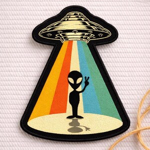 May include: A black and yellow UFO patch with a retro design. The patch features a black alien figure giving a peace sign, standing in a colorful beam of light. The patch has a black border.