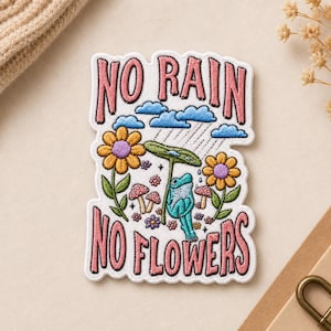 May include: An embroidered patch featuring a light blue frog holding a green leaf as an umbrella under blue clouds and rain. The design includes yellow flowers, small mushrooms, and the pink text NO RAIN NO FLOWERS on a white background.