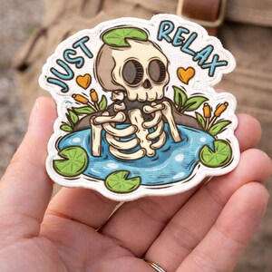 May include: A whimsical sticker featuring a skeleton relaxing in a pond with lily pads. The text "JUST RELAX" is written in a playful font. The sticker has a white border and is held in a hand.