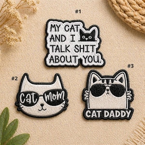 May include: Three embroidered patches with black and white cat designs. One patch reads "MY CAT AND I TALK SHIT ABOUT YOU." Another says "Cat Mom," and the third says "CAT DADDY." The patches are on a beige fabric.