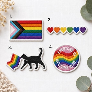 May include: A collection of embroidered patches with LGBTQ+ themes. Includes a Progress Pride flag, a row of rainbow hearts, a black cat with a rainbow flag, and a circular patch with the text "Everyone is Welcome Here."