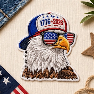 May include: An embroidered patch featuring a bald eagle wearing a blue and white baseball cap with the text 1776-2026 and sunglasses with American flag lenses. The eagle has white head feathers, a yellow beak, and brown chest feathers.