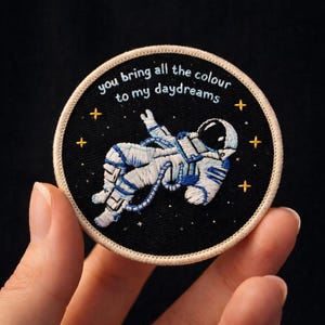 May include: Embroidered patch featuring an astronaut floating in space. The astronaut is white and blue against a black background with gold stars. The patch has the text "you bring all the colour to my daydreams" in a circular design.