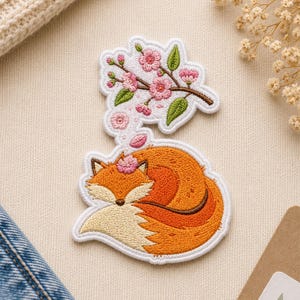 May include: Two embroidered patches: a sleeping fox with orange and cream fur, and a branch of pink cherry blossoms with green leaves. Both patches have white borders and are on a beige background.