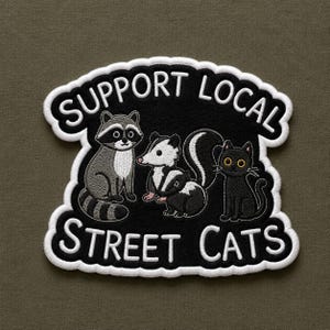 May include: An embroidered patch with a black background and white border featuring a raccoon, a skunk, and a black cat. The text SUPPORT LOCAL STREET CATS is stitched in white capital letters above and below the illustrated animals.