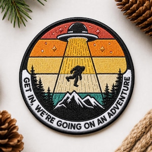 May include: Round patch featuring a retro design with a UFO beaming up a Bigfoot figure. The patch has a black border with the text "GET IN, WE'RE GOING ON AN ADVENTURE". The background has orange, yellow, and teal stripes.
