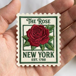 May include: A detailed, embroidered stamp featuring a red rose with green leaves. The text "The Rose" and "New York EST. 1788" are displayed in a green border. The stamp has a cream-colored background with a perforated edge.
