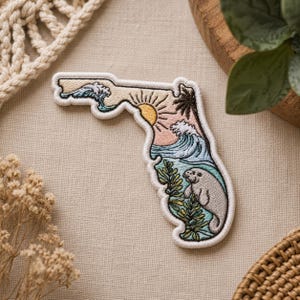 May include: Embroidered patch in the shape of Florida, depicting a beach scene with waves, a sun, palm trees, and a manatee. The patch has a white border and is on a beige background.