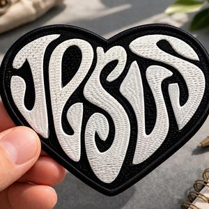 May include: Black heart-shaped patch with the word "JESUS" embroidered in white, stylized lettering. The patch has a black border and is likely an iron-on or sew-on patch for clothing or accessories.