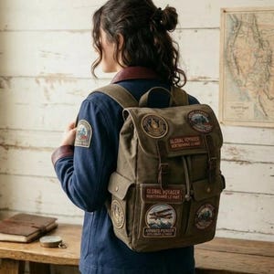 May include: A canvas backpack with multiple patches, including "Global Voyager" and an airplane design. The backpack is olive green with brown leather accents and a drawstring closure. The person is wearing a blue jacket with a patch on the sleeve.
