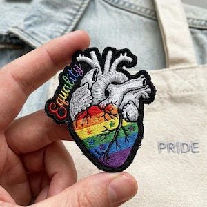 May include: Embroidered patch featuring a detailed anatomical heart design with a rainbow gradient at the bottom. The word "equality" is stitched in colorful thread. The patch is held in a hand, with a denim jacket and a tote bag with the word "PRIDE" in the background.