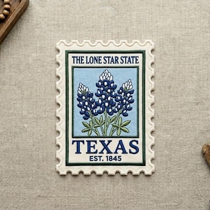 May include: Embroidered patch in the shape of a postage stamp featuring bluebonnets, the state flower of Texas. The text "THE LONE STAR STATE" and "TEXAS EST. 1845" are included in the design. The patch has a cream-colored border.