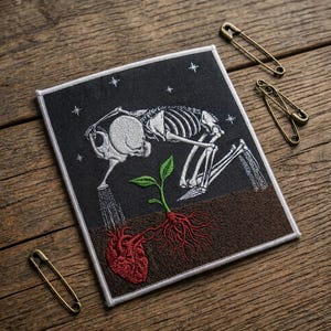 May include: Square embroidered patch featuring a skeleton watering a heart with a watering can, with a plant growing from the heart. The patch has a black background with white stars and a white border. Three gold safety pins are on a wooden surface.