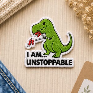 May include: Embroidered patch featuring a green dinosaur holding two red game controllers. The patch has the text "I AM... UNSTOPPABLE" in black letters on a white background. The dinosaur has a white outline.