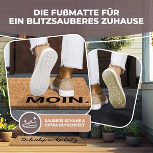 May include: A brown doormat with the word "MOIN." printed in black. The doormat has a light brown background and a dark brown border. The doormat is placed in front of a door with a white frame. The doormat is also labeled "Schuhparkplatz" in black text. The doormat is designed to help keep shoes clean and prevent slipping.