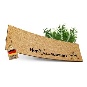 Könnte beinhalten: Eine braune Kork-Fußmatte mit dem Text "Her Wein spaziert" und einer Grafik von zwei anstoßenden Weingläsern. Ein braunes Etikett mit der deutschen Flagge und dem Text "Deutsche Marke" ist an der Fußmatte befestigt.