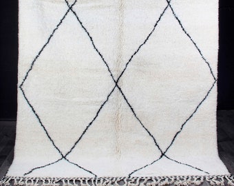 Könnte beinhalten: Ein großer, cremefarbener Teppich mit einem geometrischen Rautenmuster in Schwarz. Der Teppich hat einen dicken, strukturierten Flor und einen Fransenbesatz am unteren Rand. Der Teppich hängt vor einem dunklen Hintergrund.