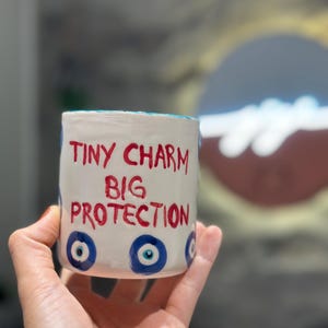 Könnte beinhalten: Ein weißer Keramikbecher mit dem handgemalten roten Text "TINY CHARM BIG PROTECTION" auf der Vorderseite. Der Becher ist mit mehreren blau-weißen Schutzaugen-Symbolen verziert, die um die Basis herum auf die glatte, zylindrische Oberfläche gemalt sind.