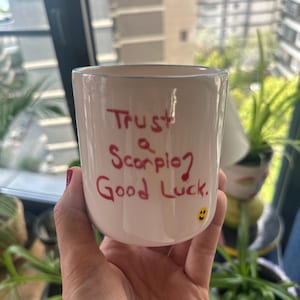 Può includere: Una tazza in ceramica bianca con testo rosso scritto a mano che recita Trust a Scorpio? Good Luck. Una piccola icona gialla con una faccina sorridente è dipinta vicino alla parte inferiore destra della tazza. La tazza ha una forma cilindrica.