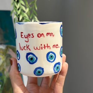 May include: A white ceramic mug featuring hand-painted blue and turquoise evil eye symbols scattered across the surface. The text "Eyes on me, luck with me." is written in red paint on the front. The mug has a simple cylindrical shape.