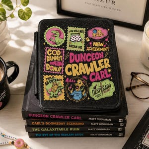 May include: A black e-reader case featuring colorful stickers with text including Dungeon Crawler Carl, God Damnit Donut, and Desperado. The case rests on a stack of four books by Matt Dinniman, including titles like The Eye of the Bedlam Bride.