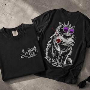 May include: A black t-shirt featuring a small white graphic on the front that reads The Desperado Club So Fun It Hurts. The back displays a large white line art illustration of a fluffy cat wearing a crown and purple sunglasses, holding a martini glass with a red cherry.