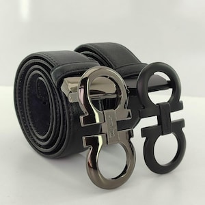 May include: Two black leather belts with silver and black buckles. The buckles feature a distinctive, interlocking design. One belt is rolled up, while the other is partially unrolled, showcasing the textured leather and buckle details. The background is plain white.
