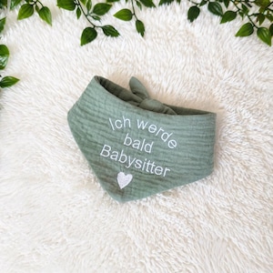 Könnte beinhalten: Ein dreieckiges Bandana-Lätzchen aus salbeigrünem, strukturiertem Stoff. Das Lätzchen ist mit dem weißen Sticktext Ich werde bald Babysitter und einem kleinen weißen Herzsymbol unter dem Text versehen. Es verfügt über einen Bindeknoten am oberen Rand.