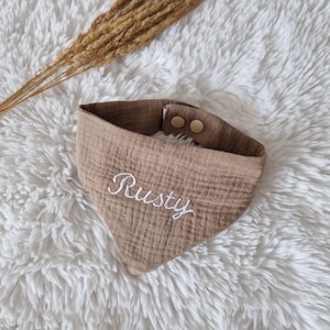 Könnte beinhalten: Ein braunes Hunde-Bandana mit dem Namen "Rusty" in weißer Schrift gestickt. Das Bandana hat eine dreieckige Form und wird mit zwei Druckknöpfen befestigt. Der Hintergrund ist eine flauschige weiße Oberfläche, mit getrockneten Weizenähren.