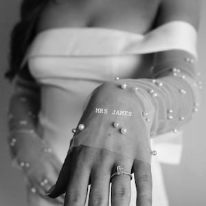 May include: A bride's hand with a diamond ring, wearing a white off-the-shoulder dress with a sheer beaded sleeve that says "MRS. JAMES".