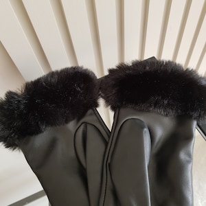 May include: A pair of black gloves featuring a smooth, matte finish on the main body and a thick, plush black faux fur trim around the wrist openings. The gloves are positioned side-by-side, showcasing the sleek texture and the soft fur cuffs.