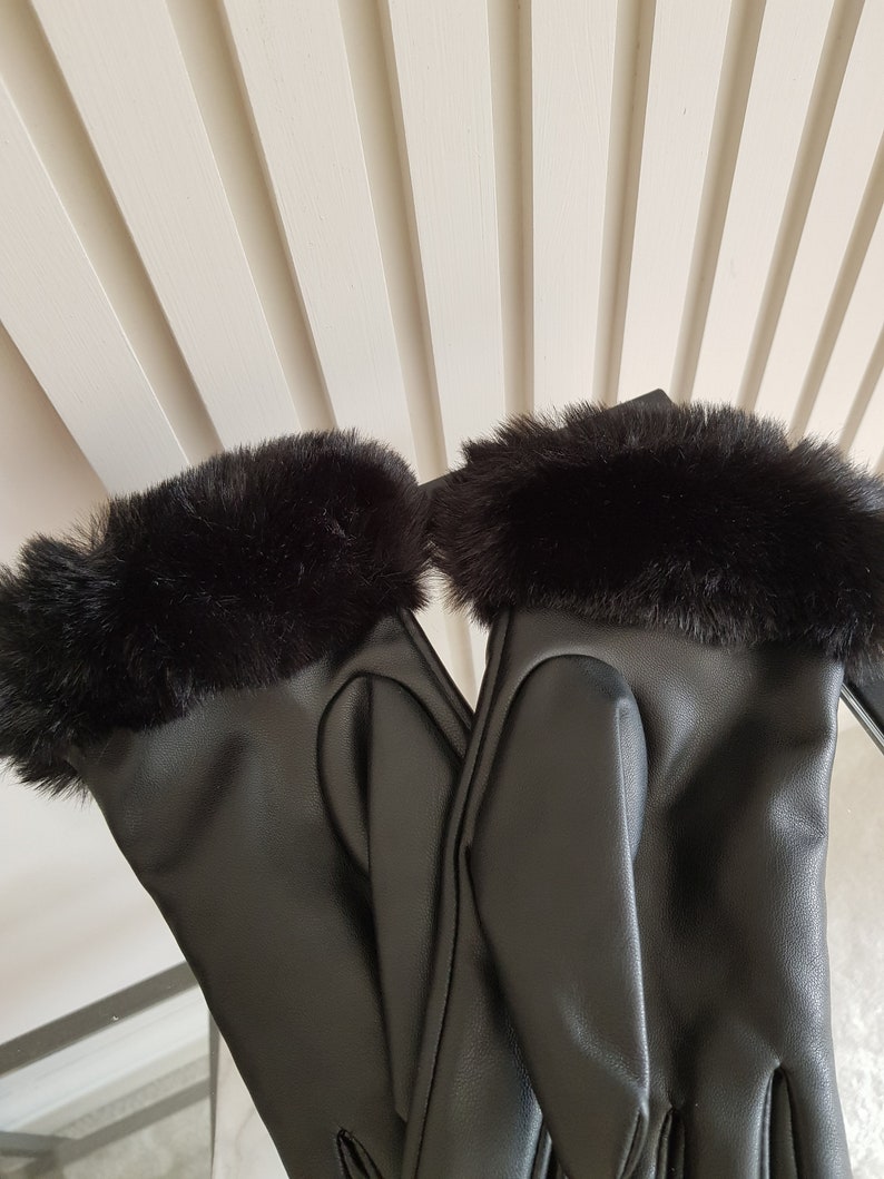 May include: A pair of black gloves featuring a smooth, matte finish on the main body and a thick, plush black faux fur trim around the wrist openings. The gloves are positioned side-by-side, showcasing the sleek texture and the soft fur cuffs.