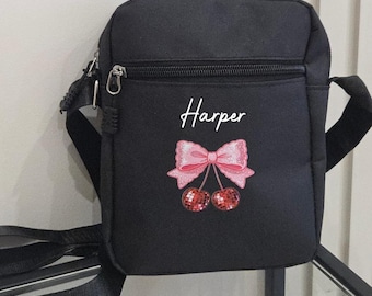 May include: Black crossbody bag with a zippered front pocket and adjustable strap. The bag features the name "Harper" in white script above an embroidered design of a pink bow and two red cherries. The bag is made of a durable material.