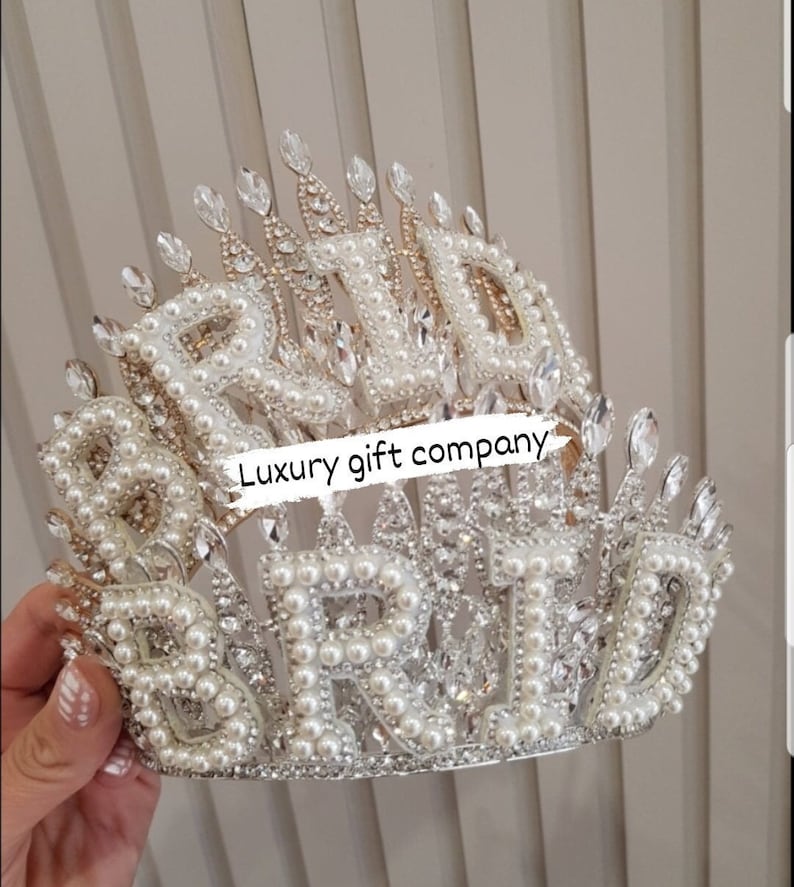 May include: A silver and white crown with the word "BRIDE" spelled out in large, pearl-encrusted letters. The crown is decorated with rhinestones and has a pointed top.