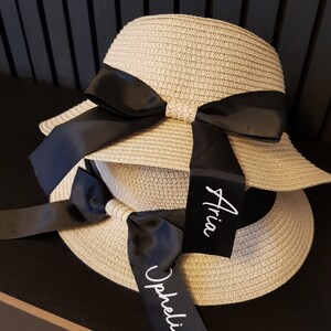 May include: Two straw sun hats with black ribbon bows and personalized tags. The tags are labeled "Aria" and "Ophelia".