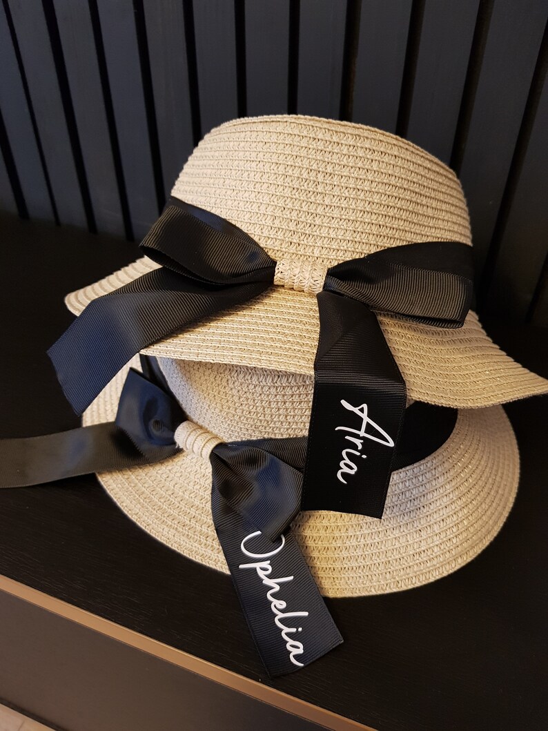 May include: Two straw sun hats with black ribbon bows and personalized tags. The tags are labeled "Aria" and "Ophelia".