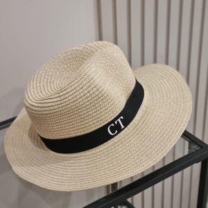 May include: A beige straw hat with a black ribbon and the initials "CT" embroidered in white.