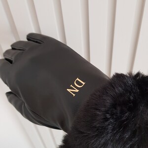 May include: A black glove featuring a smooth finish and a plush black faux fur cuff at the wrist. The back of the glove is personalised with the gold-coloured capital letters DN, which are positioned vertically in a clean, sans-serif font.