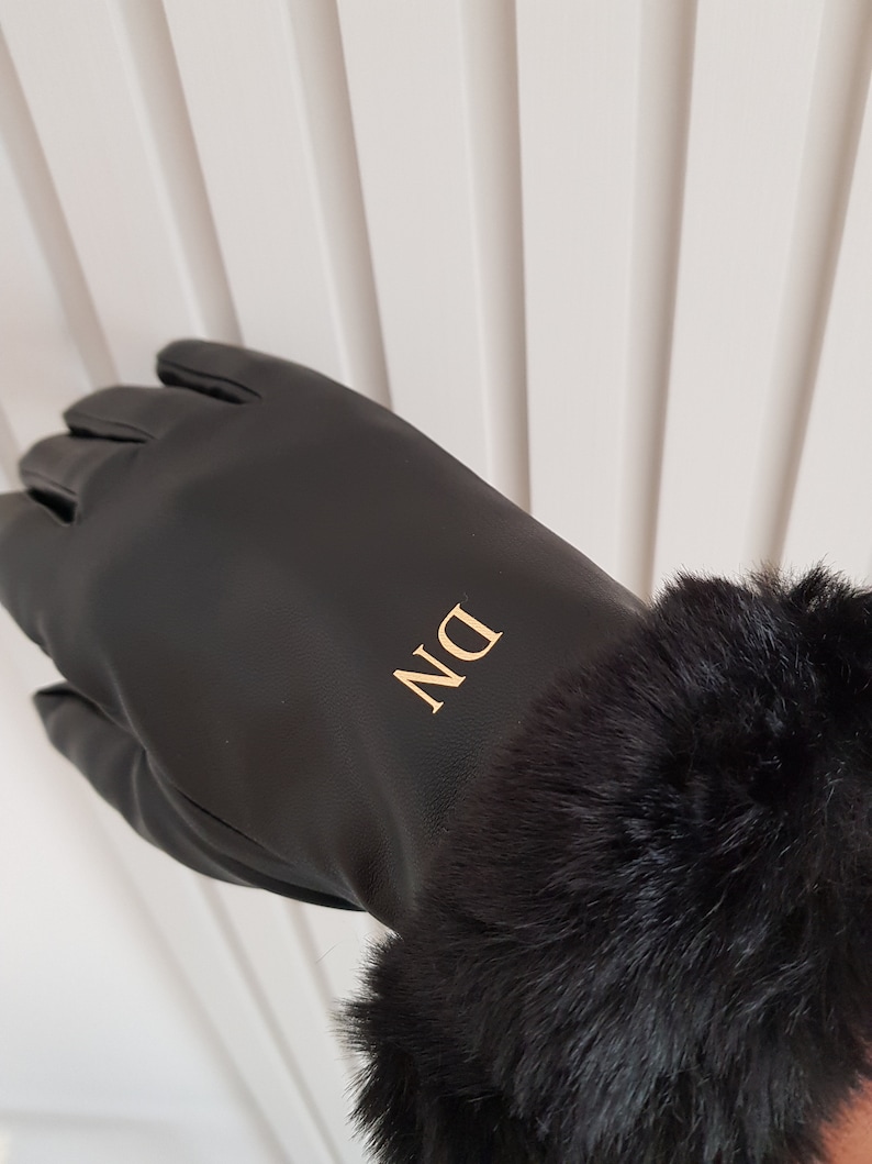 May include: A black glove featuring a smooth finish and a plush black faux fur cuff at the wrist. The back of the glove is personalised with the gold-coloured capital letters DN, which are positioned vertically in a clean, sans-serif font.